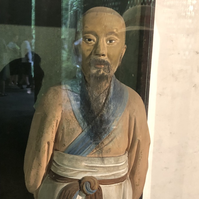 A painted clay figure of a bearded sage behind glass