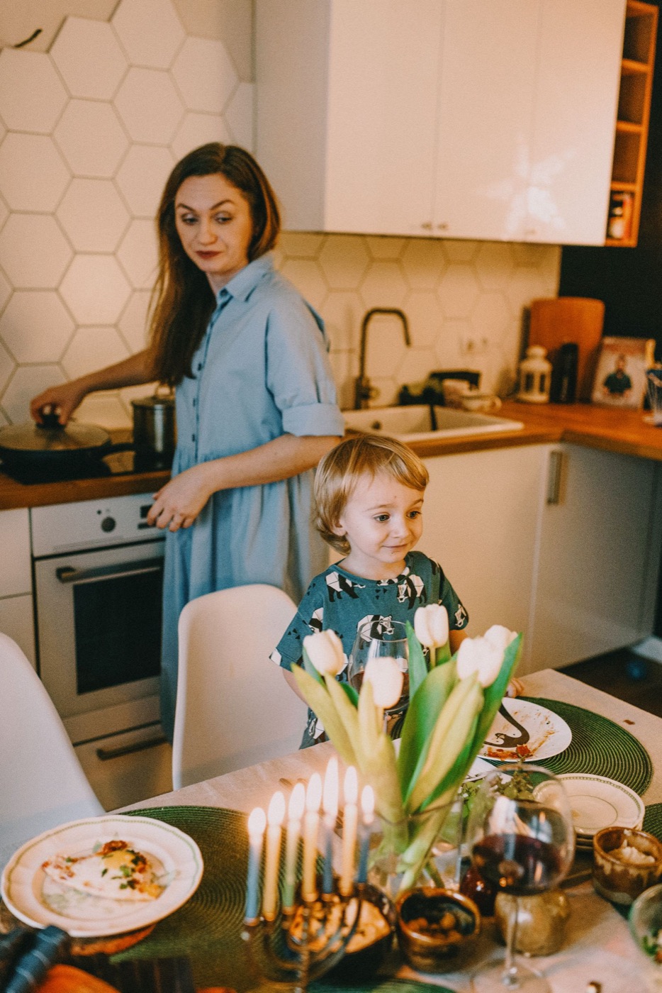 A mother at the stove in a warm kitchen, a child at the table with lit candles